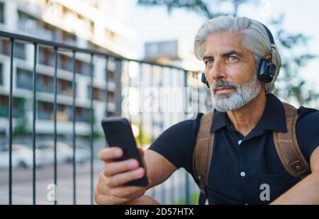 Porträt eines reifen Mannes mit Kopfhörern, die im Freien in der Stadt sitzen, mit dem Smartphone. Stockfoto
