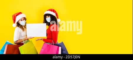 Zwei junge niedliche Frauen tragen Weihnachtsmann Hut Blick auf die Kamera mit breiten Augen und medizinische Maske, halten Schild für Kopie Platz mit vielen Einkaufstasche Stockfoto