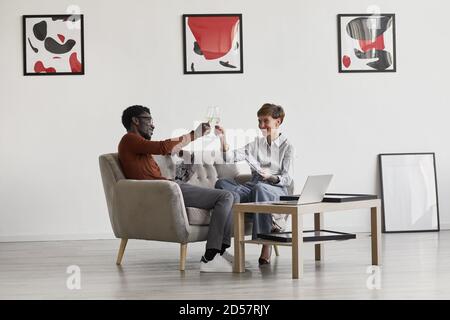 Porträt eines jungen Mischrennpaares, das Champagner genießt, während es auf der Couch im Zimmer sitzt, das mit moderner Kunst dekoriert ist, Kopierraum Stockfoto