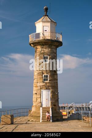 Ein Tourist schaut auf ein Telefon, während er auf Stein sitzt Schritte am Fuße des historischen Sandsteins Whitby West Pier Leuchtturm an einem sonnigen Nachmittag Stockfoto