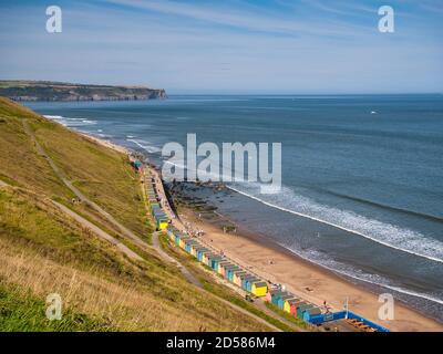 Die Küste nördlich von Whitby, North Yorkshire, UK an einem klaren sonnigen Tag im Sommer, mit bunten Strandhütten im Vordergrund Stockfoto