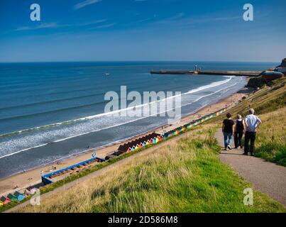 Touristen auf einem Pfad mit Blick auf bunte Strandhütten, die Küste und Hafen Piers in Whitby, North Yorkshire, Großbritannien Stockfoto