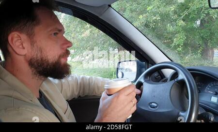 Ein bärtiger Kaukasischer Mann mittleren Alters, der mit einem Auto durch die Stadt fährt und Kaffee trinkt. Stockfoto