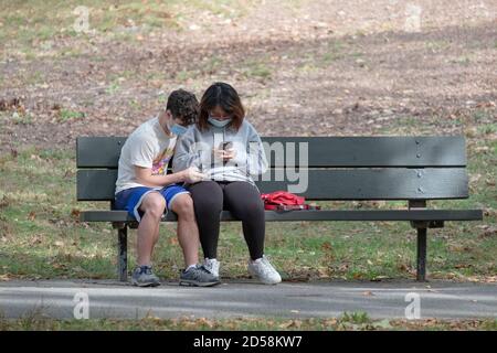 Ein junges Paar sitzt zusammen auf einer Parkbank, jedes mit seinem eigenen Handy eingetaucht. In Flushing, Queens, New York City.trägt, trägt Stockfoto
