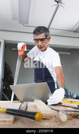 Senior man Hold sah und überprüfen sein Projekt in Laptop-Computer in der Schreinerei Werkstatt. Konzept Senioren Hobby nach dem Ruhestand. Stockfoto