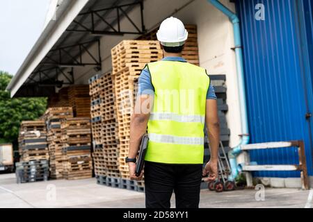 Porträt von jungen Arbeiter arbeiten in der Logistikbranche im Freien vor dem Fabriklager. Mann mit Hut, der die Kamera beim Gehen anschaut und einen Laptop am Depot mit Palettenstapel hält Stockfoto