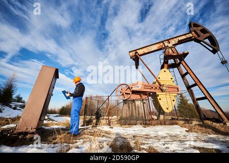 Petroleum Ingenieur in Arbeit Overalls und Helm Notizen auf Zwischenablage während der Steuerung der Arbeit der Ölpumpeinheit unter schönem Himmel. Konzept der Ölförderung und Erdölindustrie. Stockfoto