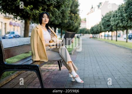 Junge hübsche intelligente konzentrierte asiatische Geschäftsfrau, in eleganten bunten gelb beige Anzug, sitzt auf der Stadt Bank mit ihrem Laptop. Kostenloses Wi-Fi in Stockfoto