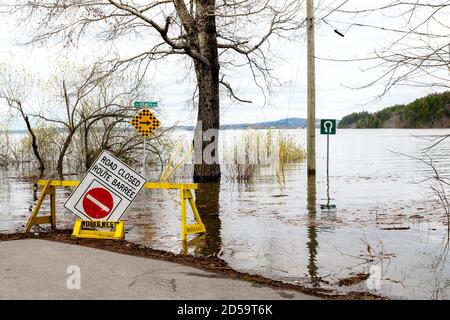 Eine überflutete Straße. Eine Barrikade und ein Schild "Straße geschlossen" blockieren den Weg. Nach der Barrikade ist die Straße komplett überflutet. Bäume und ein Mast umgeben Stockfoto