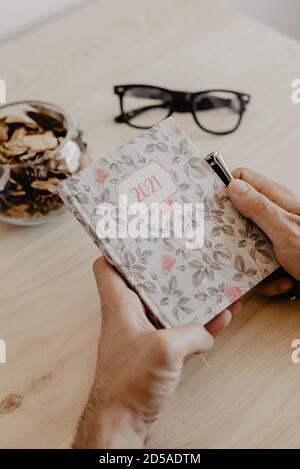 Ein Mann, der an einem Schreibtisch sitzt, plant das Jahr 2021 Auf einem Tagebuch mit einem Stift in der Hand und Brille an seiner Seite Stockfoto