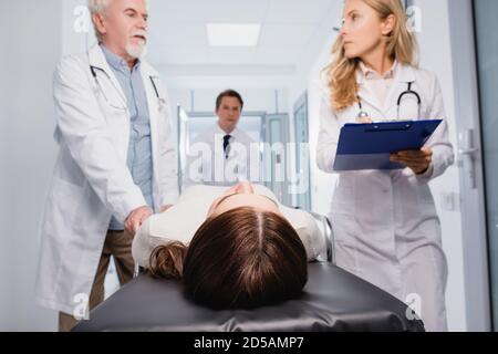 Selektiver Fokus der kranken Frau, die auf einer Bahre in der Nähe von Ärzten liegt Mit Zwischenablage in der Klinik Stockfoto