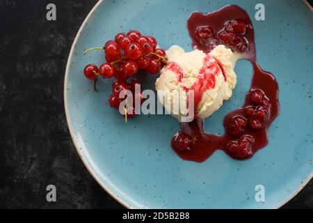 Vanilleeis-Schaufel mit roten Johannisbeeren und Obstsauce auf blauem Teller, dunkler Hintergrund, Kopierraum, Hochwinkelansicht von oben, ausgewählter Fokus Stockfoto