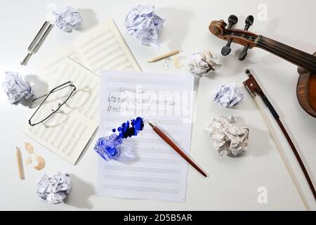 Der Arbeitsplatz des Musikers, die zusammengeknitterten und flachen Notenblätter, die handschriftliche Notation der musikalischen Komposition und das umgedrehte Tintenfass auf dem hellgrauen Register Stockfoto