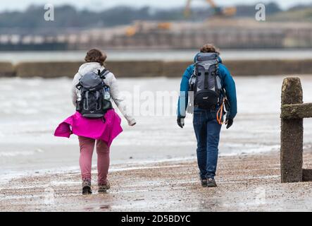 Pärchen, die bei nassem und windigem Wetter in Großbritannien an einem Strand mit Rucksäcken (Rucksäcken) spazieren. Pärchen auf Herbstspaziergang, jeder mit Rucksack (Rucksack). Stockfoto