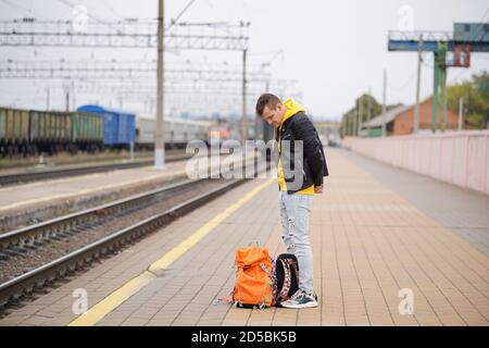 Der junge Mann steht auf dem Bahnsteig und wartet auf den Zug. Männlicher Passagier mit Rucksäcken auf Bahnsteig in Warten auf Zugfahrt. Konzept des Tourismus Stockfoto