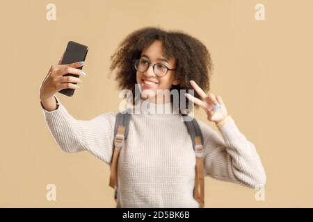 Selfie für Blog und zurück zur Schule. Lächelnde afroamerikanische junge Frau in Brille mit Rucksack zeigt Frieden Stockfoto