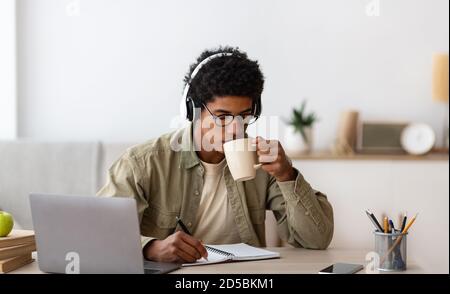 Afroamerikanischer Teenager mit Kopfhörern, der fernab von zu Hause studiert, Kaffee trinkt und Notizen im Copybook macht Stockfoto