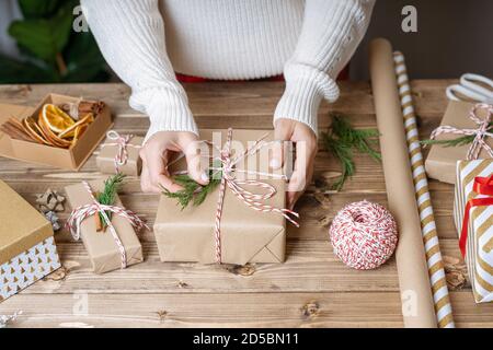 Frau s Hände Verpackung Weihnachtsgeschenk, Nahaufnahme. Unvorbereitete Geschenke auf Holzhintergrund mit Dekorelementen und Gegenständen, Draufsicht. Weihnachten oder Neu Stockfoto