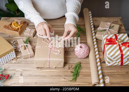 Frau s Hände wickeln Weihnachtsgeschenk, aus der Nähe. Unvorbereitet Weihnachtsgeschenke auf Holzuntergrund mit Dekor Elemente und Elemente, Ansicht von oben. Weihnachten Stockfoto