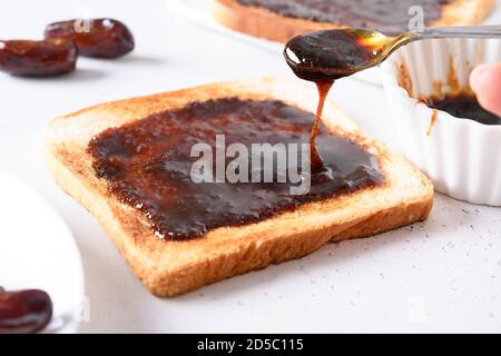 Frau Gießen Datteln Marmelade auf frischen knusprigen Toast auf weiß. Nahaufnahme. Stockfoto