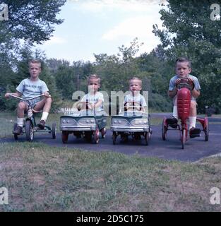 1960, historisch, draußen auf Asphalt bei einem Feld, vier kleine Jungen, Brüder und alle tragen passende hellblaue T-Shirts und Shorts, sitzen auf ihren Metall-Ride-on-Spielzeugautos und Dreiräder der Zeit. In den 1950er und 60er Jahren war AMF Junior einer der größten Hersteller von Kinderspielzeug. Stockfoto