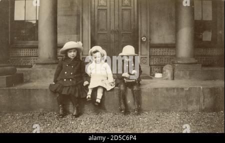 WW! Foto der Ära Anfang des 20. Jahrhunderts von 3 kleinen Kindern, die Hüte tragen, die auf den Stufen des Grand House sitzen, um 1915, einer von ihnen schaut auf eine Kamera die Kinder tragen zweireihige Mäntel und Kutten schützen ihre Schuhe, Großbritannien Stockfoto