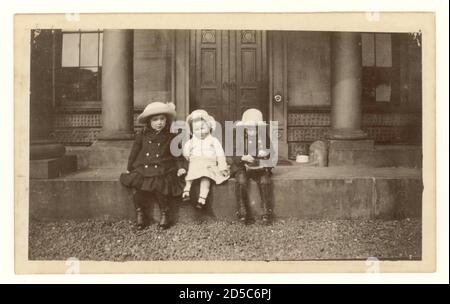 WW1 Ära Anfang 1900 Foto von 3 kleinen Kindern tragen Hüte sitzen auf den Stufen des Grand House, um 1915, einer von ihnen ist auf eine Kamera die Kinder tragen zweireihige Mäntel und Kutten schützen ihre Schuhe, UK Stockfoto