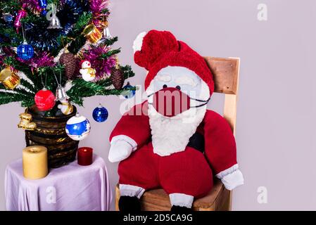 Foto eines gefertigten Weihnachtsmanns in einem chirurgischen Rot Maske sitzt auf einem kleinen Holzstuhl neben einem Weihnachtsbaum Stockfoto