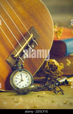 Eine antike Taschenuhr lehnte sich an eine Ukulele und ein altes Buch mit Vintage-Karte und Messingstift auf Holztisch platziert. Nahaufnahme und Kopie Platz für Text. Stockfoto