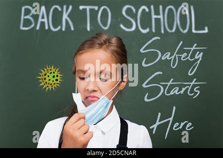 Selektiver Fokus der unzufrieden Schulmädchen berühren Schutzmaske in der Nähe Kreidetafel Mit Sicherheit beginnt hier Schriftzug im Klassenzimmer Stockfoto