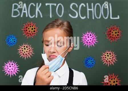Selektiver Fokus der unzufrieden Schulmädchen berühren Schutzmaske in der Nähe Kreidetafel Mit Schulschrift und Virusillustration Stockfoto