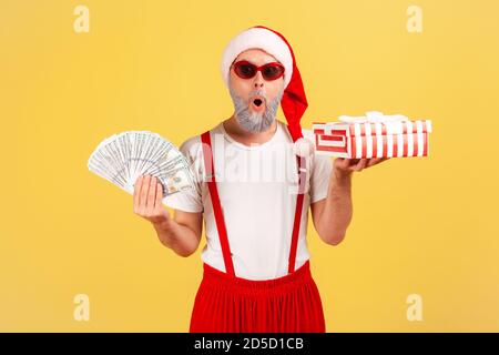 Aufgeregt überrascht älteren Mann in weihnachtsmann Hut und stilvolle Sonnenbrille hält geschenkbox und Dollar-Banknoten, Urlaub Boni. Innenaufnahmen im Studio Stockfoto