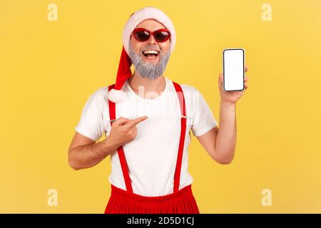 Glücklich lächelnder Mann mit grauem Bart in Sonnenbrille und weihnachtsmann Hut zeigt Finger auf Smartphone mit leerem weißen Display, Online-Anwendung. Innen Stockfoto
