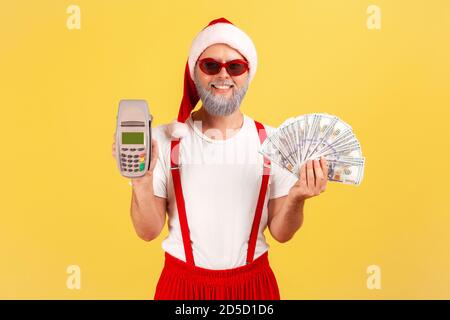 Positive Erwachsene Mann in Sonnenbrille und weihnachtsmann Hut hält POS Terminal und Dollar-Banknoten lächeln an der Kamera, wählen bargeldlose Zahlungen. Innen s Stockfoto