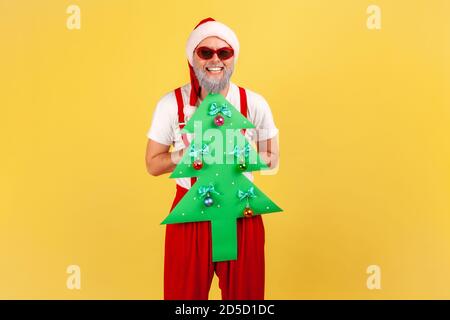 Extrem glücklicher älterer Mann im weihnachtsmann Kostüm und stilvolle Sonnenbrille hält Papier weihnachtsbaum Blick auf die Kamera mit toothy Lächeln. Stu Stockfoto