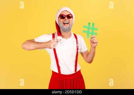 Glücklich lächelnder graubärtiger Mann in Sonnenbrille und weihnachtsmannhut, der auf grünes Hashtag-Symbol in seiner Hand zeigt und Feiertagsposten markiert. Studio-Zimmer im Innenbereich Stockfoto