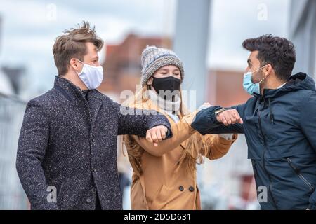 Zwei Männer und eine Frau grüßen sich mit dem Ellenbogen. Corona-Zeit mit alltäglichen Masken in der kalten Jahreszeit in der Stadt. Stockfoto
