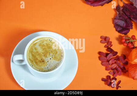 Weiße Tasse Kaffee mit Cappuccino-Creme auf orangefarbenem Hintergrund mit Herbstblättern, Äpfeln und roten Beeren. Herbstsaison, Freizeit und Kaffee Stockfoto
