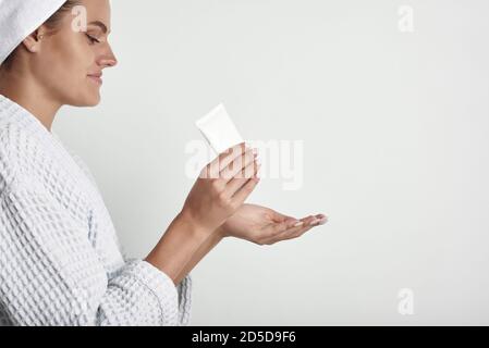 Eine junge Frau in einem Bademantel und mit einem Handtuch auf dem Kopf drückt sich Handcreme aus. Frauenhände mit Vitiligo. Stockfoto