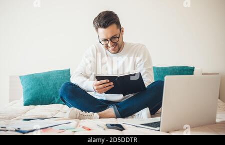 Der junge kaukasische Unternehmer lächelt und arbeitet mit einigen Papieren Zu Hause sitzen auf dem Sofa und mit Laptop Stockfoto