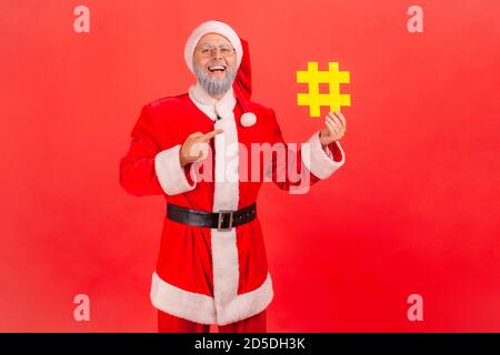 Positive glücklich grauen bärtigen Mann in Brillen und weihnachtsmann Kostüm zeigt Finger auf gelbe Hashtag-Symbol in der Hand, Tagging Feiertagsbeiträge. Ind Stockfoto