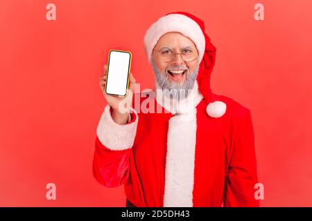 Glücklich grau bärtig weihnachtsmann hält und zeigt Smartphone mit weißem Display Blick auf die Kamera mit toothy Lächeln. Innenaufnahme des Studios isoliert auf r Stockfoto