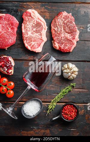 Verschiedene rohe Rindersteaks mit Gewürzen und Rotwein in Flasche und Glas auf alten dunklen Holzplanken Draufsicht. Stockfoto