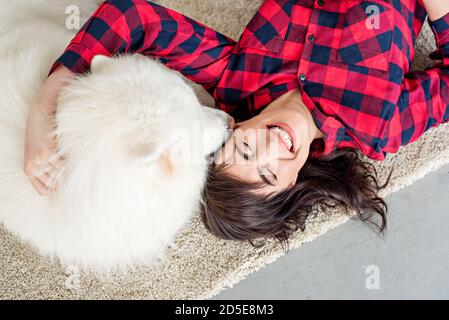 Schöne Brünette Mädchen mit Locken in einem rot karierten Hemd lacht umarmt eine samoyed Husky, die das Mädchen auf die Wange leckt. Hintergrund eines Weihnachtsbaums Stockfoto