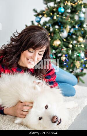 Schöne Brünette Mädchen mit Locken in einem rot karierten Hemd lacht umarmt eine samoyed Husky, die das Mädchen auf die Wange leckt. Hintergrund eines Weihnachtsbaums Stockfoto