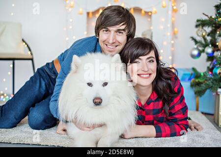 Junger Mann mit einer schönen Frau umarmt einen weißen samoyed Hund. Nahaufnahme Porträt einer glücklichen Familie mit einem Samodes laika in der Nähe eines weißen Kamins mit Girlande Stockfoto