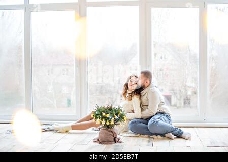 Glückliches verliebtes Paar sitzt auf dem Holzboden am Panoramafenster. Ein junger Mann in einem weißen Pullover mit einem Bart umarmt, küsst eine schöne Frau Stockfoto