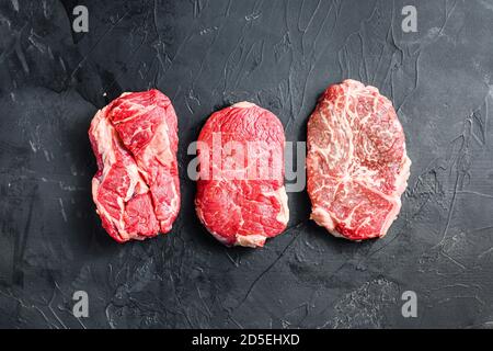 Set von rohen alternativen Schnitten Rindersteaks, obere Klinge, Rumpf und Futterrolle, auf schwarzem texturierten Hintergrund, Draufsicht. Stockfoto