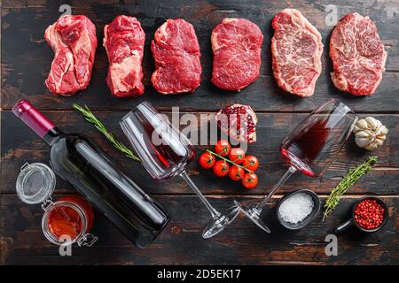 Verschiedene rohe Rindersteaks mit Gewürzen und Rotwein in Flasche und Glas auf alten dunklen Holzplanken Draufsicht. Stockfoto