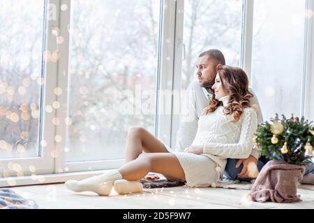 Glückliches verliebtes Paar sitzt auf dem Holzboden am Panoramafenster. Ein junger Mann in einem weißen Pullover mit einem Bart umarmt, küsst eine schöne Frau Stockfoto